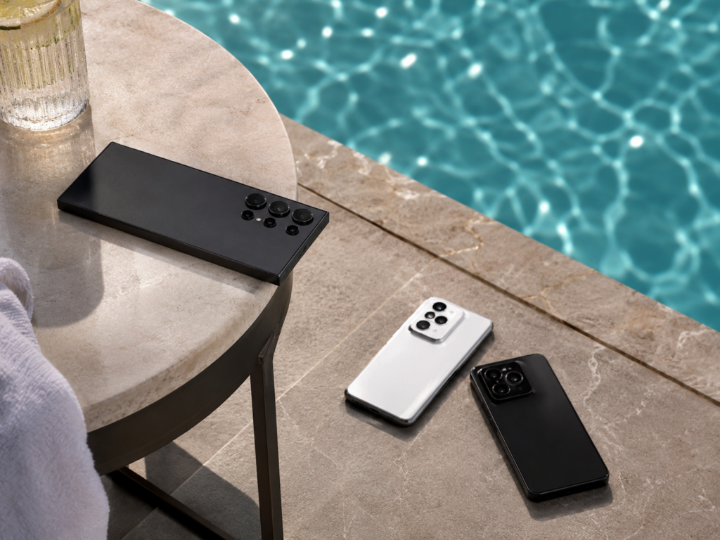 Três celulares resistentes em cena ao lado da piscina: um Samsung Galaxy S26 Ultra sobre a mesa próxima à borda e, no chão, um Motorola Edge 70 Fusion e um Xiaomi POCO X8 Pro.